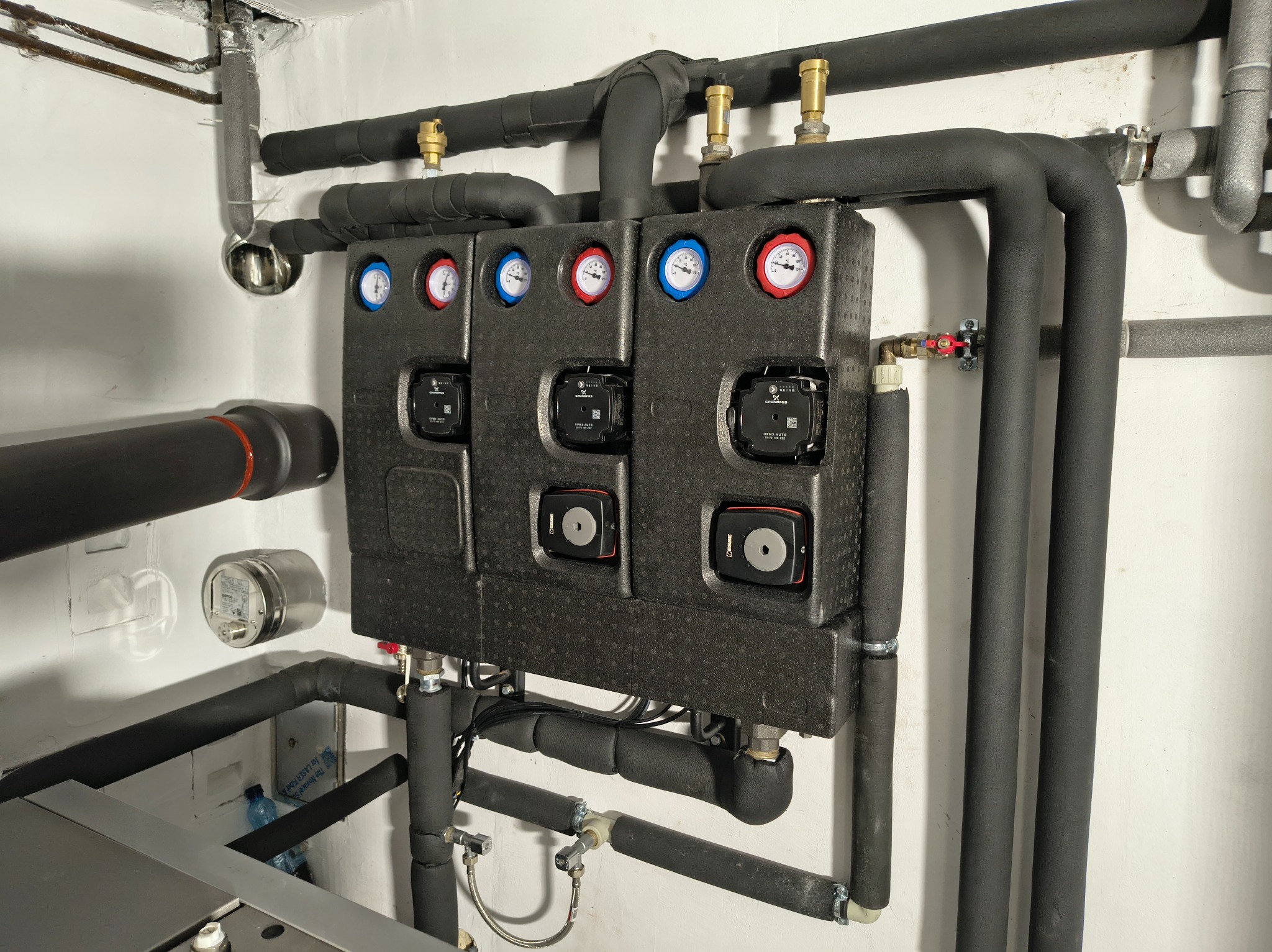 Rozdzielacz ogrzewania podłogowego z termometrami i pompami obiegowymi, zaizolowane rury w kotłowni. Widoczne elementy instalacji CO.