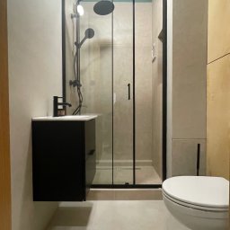 Monika Latarska - Nowoczesna łazienka z kabiną prysznicową typu walk-in z czarną ramą, wiszącą szafką z umywalką i czarną armaturą oraz podwieszaną toaletą, wykończona w odcieniach beżu i szarości z akcentem...