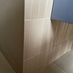 Monika Latarska - Ułożone pionowo, błyszczące, kremowe płytki typu cegiełka na ścianie łazienki, z fragmentem niebieskiej szafki wiszącej i szarą podłogą z płytek.