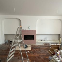 Monika Latarska - Wnętrze w trakcie remontu w Warszawie: widoczny kominek z obudową w kolorze pudrowego różu, drabina aluminiowa, częściowo położona drewniana podłoga i przygotowane materiały budowlane. Ściany...