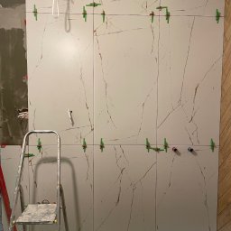 Monika Latarska - Montaż wielkoformatowych płytek imitujących marmur na ścianie, z użyciem poziomowania krzyżykami, widoczna drabina i fragment instalacji wodnej.