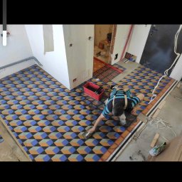 Monika Latarska - Układanie wzorzystej podłogi w pomieszczeniu, widoczny pracownik rozprowadzający fugę na nowo położonych płytkach z geometrycznym wzorem 3D, czerwone wiadro z narzędziami obok.