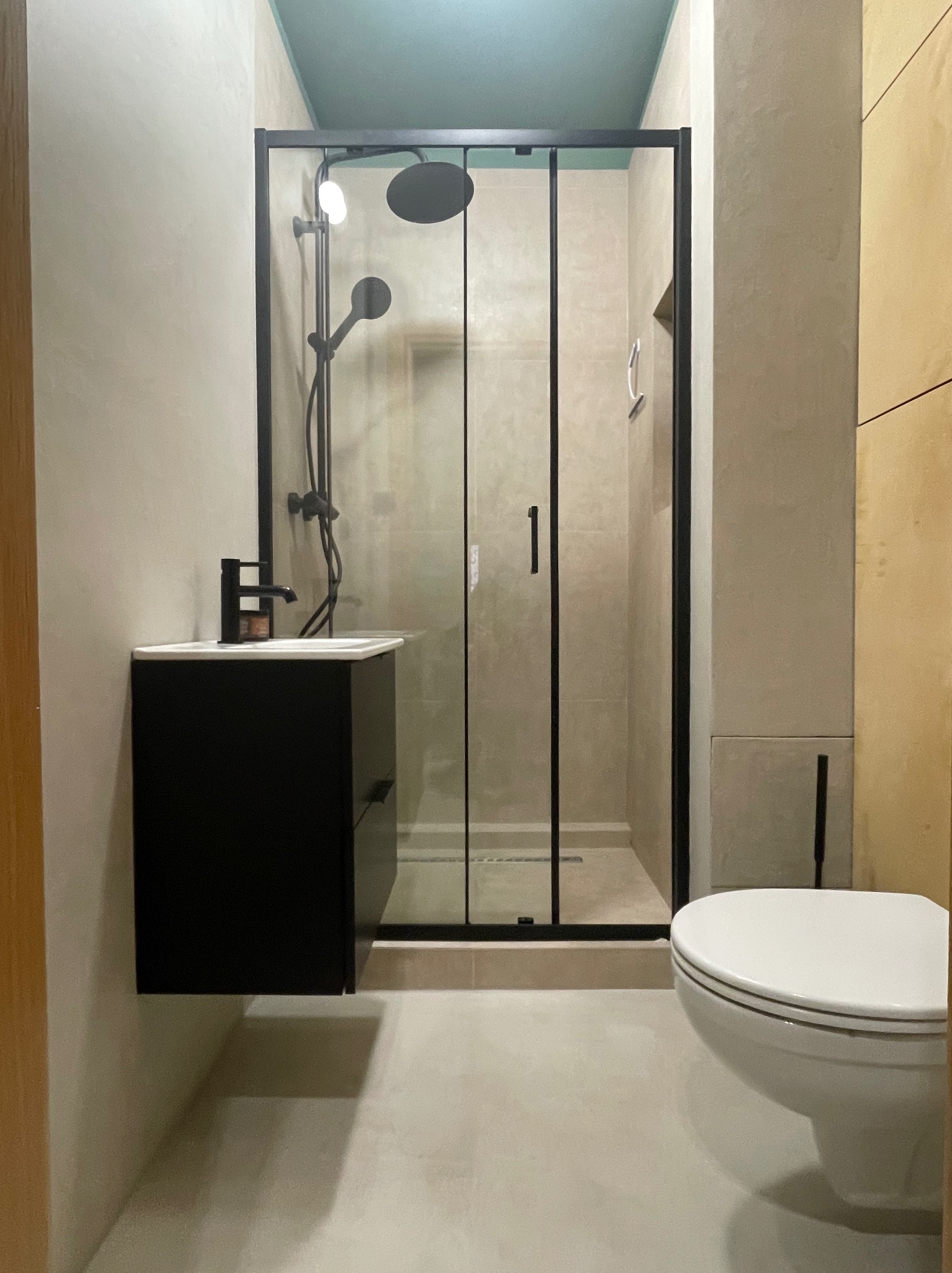 Nowoczesna łazienka z kabiną prysznicową typu walk-in z czarną ramą, wiszącą szafką z umywalką i czarną armaturą oraz podwieszaną toaletą, wykończona w odcieniach beżu i szarości z akcentem...