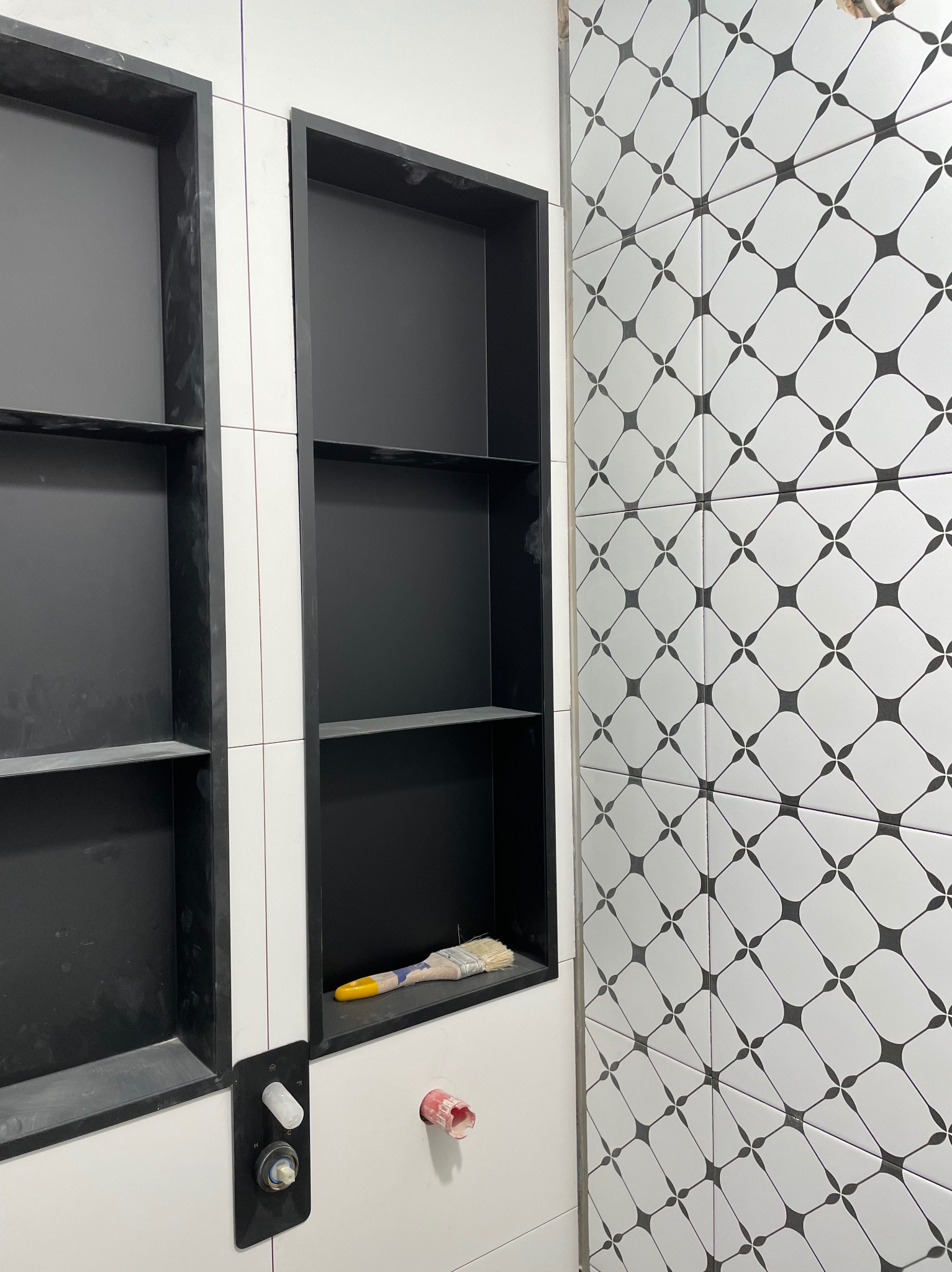 Ściana łazienki z białymi płytkami w czarne wzory, wnęki z czarnym wnętrzem, widoczny pędzel malarski i elementy instalacji wodnej.