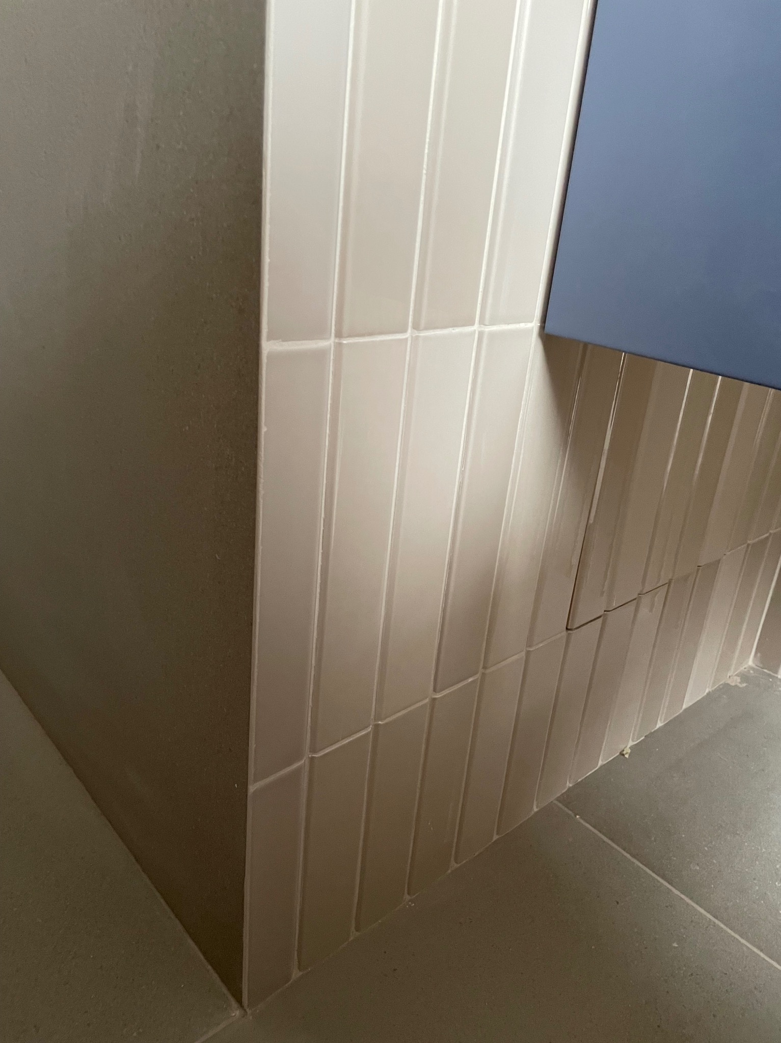 Ułożone pionowo, błyszczące, kremowe płytki typu cegiełka na ścianie łazienki, z fragmentem niebieskiej szafki wiszącej i szarą podłogą z płytek.