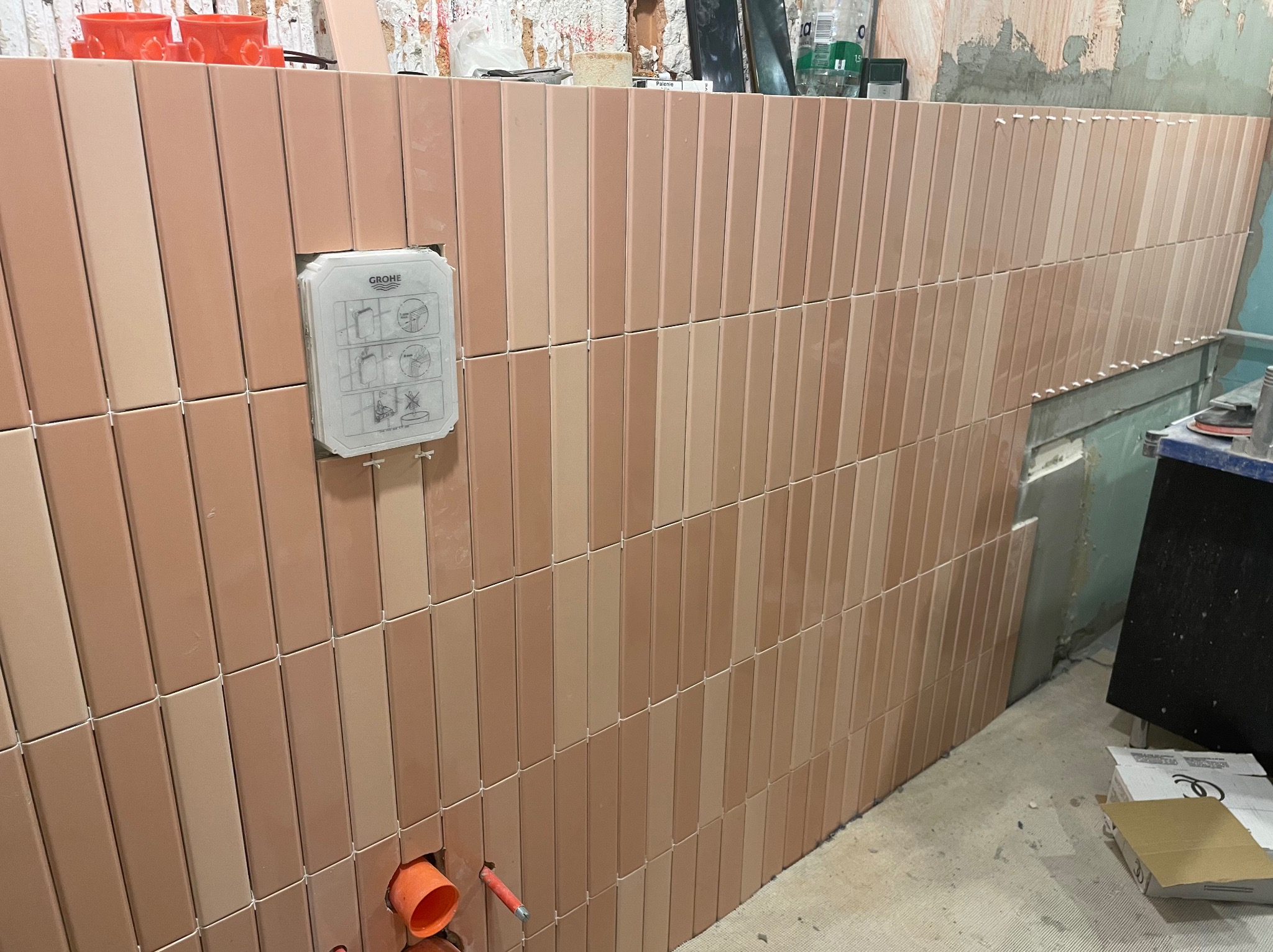 Ściana łazienki wyłożona pionowymi, różowymi płytkami w dwóch odcieniach, z zamontowaną puszką podtynkową Grohe i widocznymi elementami instalacji hydraulicznej.