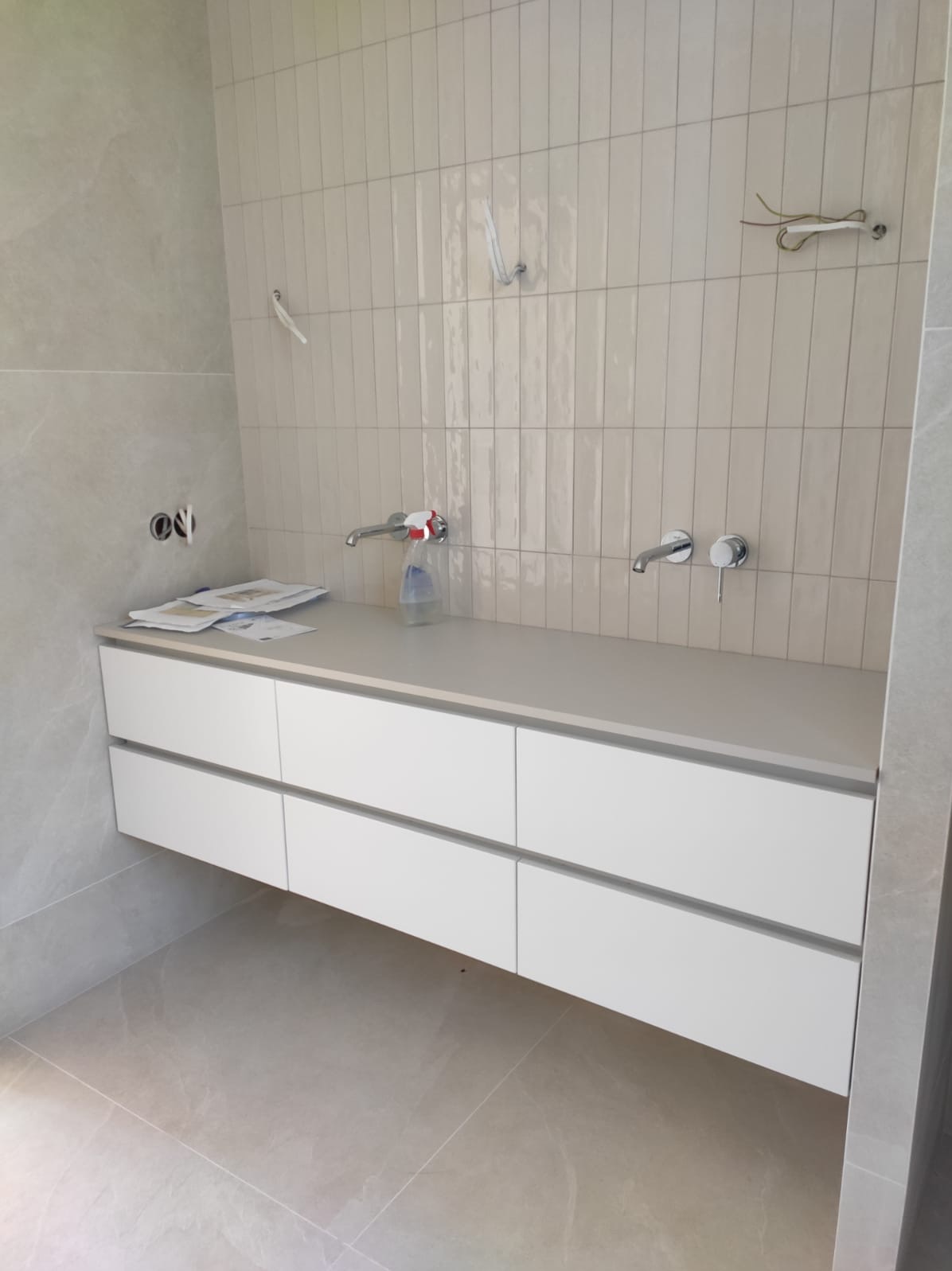 Nowoczesna łazienka w trakcie wykańczania: biała szafka pod umywalkę z sześcioma szufladami, jasne płytki na ścianie ułożone pionowo, widoczne przyłącza i akcesoria montażowe, butelka środka...