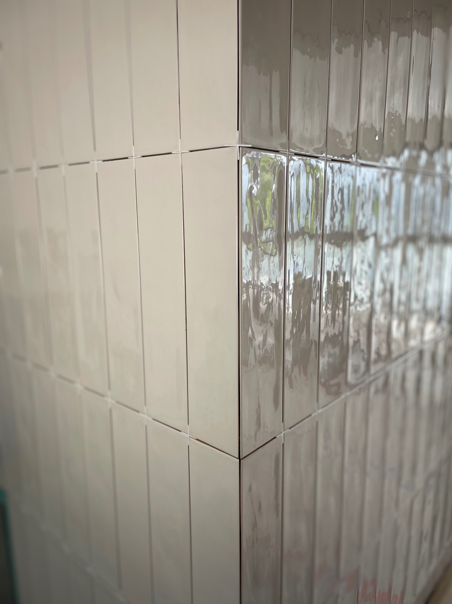 Ułożone pionowo błyszczące, beżowe płytki ceramiczne typu 'cegiełka' na ścianie, widoczny narożnik z plastikowymi krzyżykami dystansowymi.