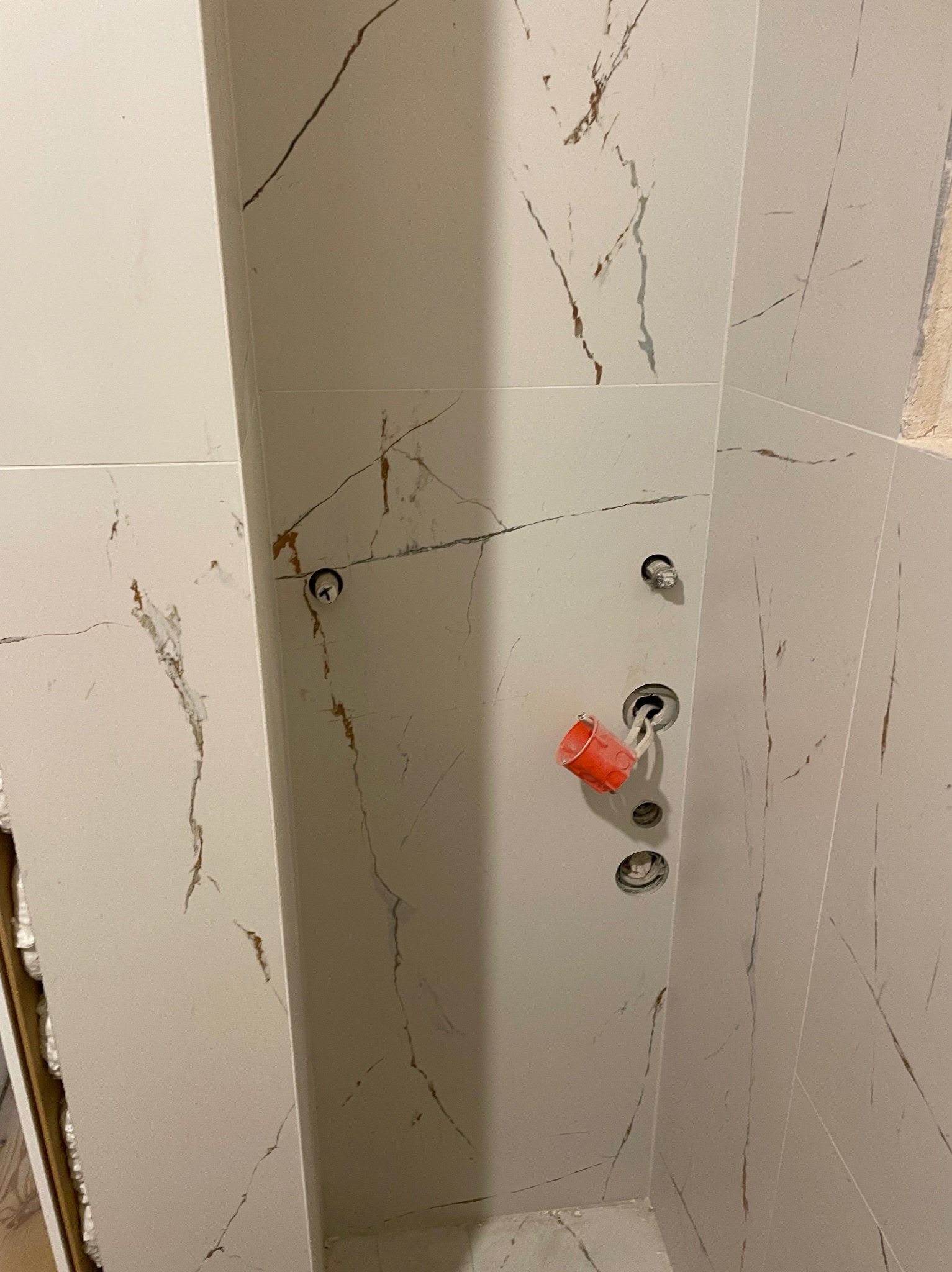 Ściana łazienki wyłożona białymi płytkami imitującymi marmur z widocznymi otworami na armaturę i zabezpieczeniem w postaci czerwonego kubka z taśmą.