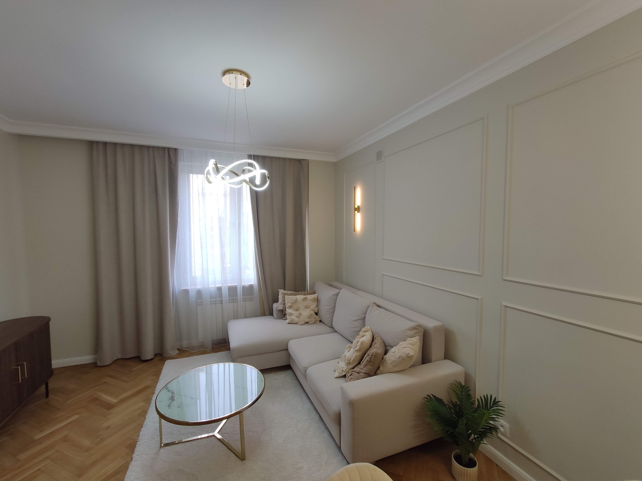 Jasny salon z parkietem w jodełkę, beżową sofą narożną, okrągłym stolikiem kawowym z marmurowym blatem i nowoczesną lampą wiszącą.