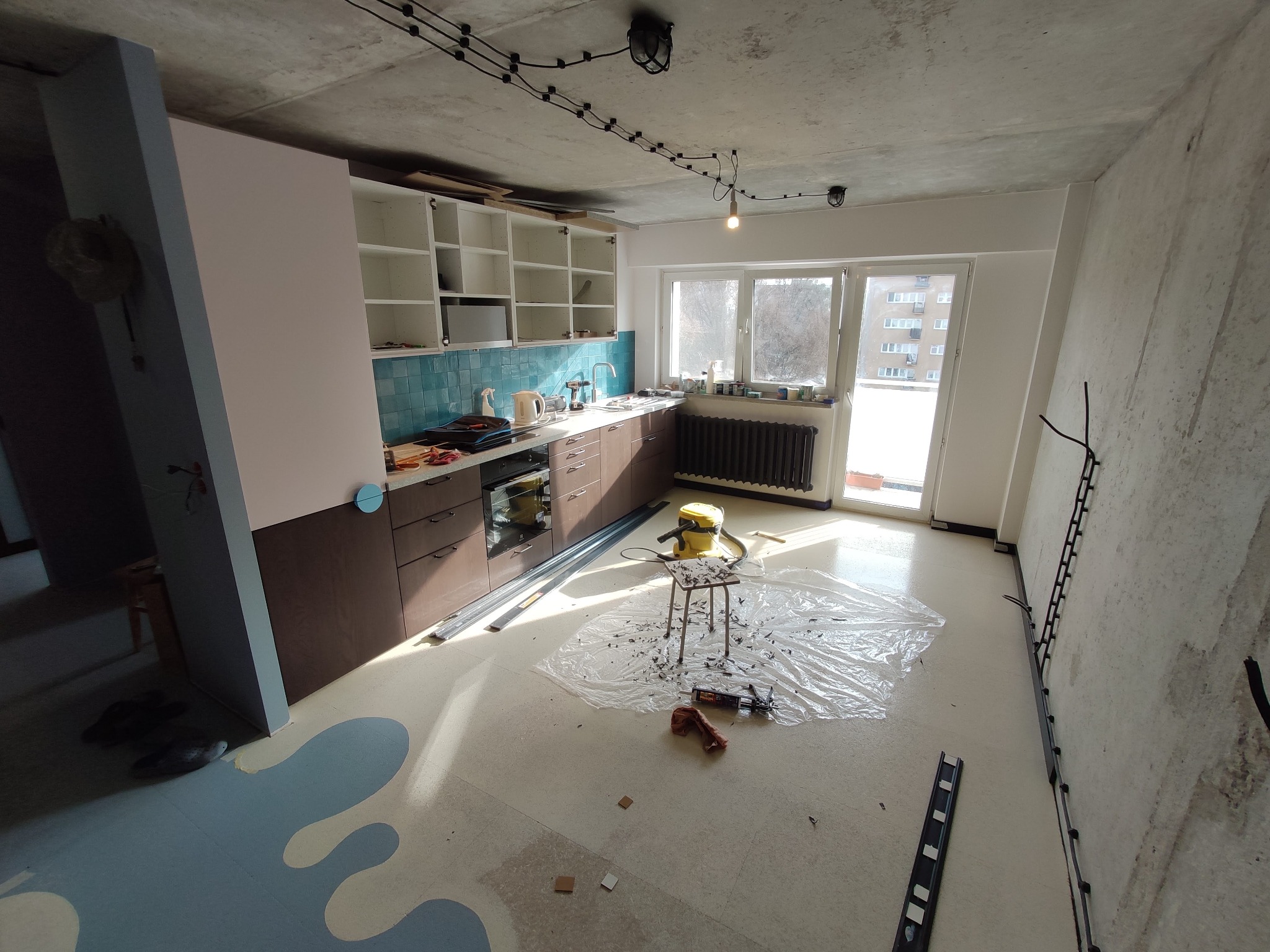 Wnętrze kuchni w trakcie prac wykończeniowych: widoczne częściowo zamontowane szafki kuchenne w kolorach brązowym i białym z turkusowymi płytkami nad blatem, narzędzia i akcesoria na podłodze...