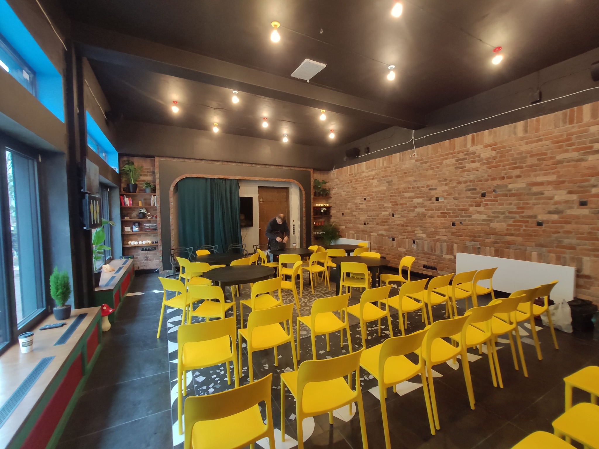 Sala konferencyjna z rzędami żółtych krzeseł i okrągłymi stolikami na tle ściany z cegły, oświetlona lampami edisona. W tle widoczna osoba. Nietypowy wystrój z elementami roślinnymi przy oknie.
