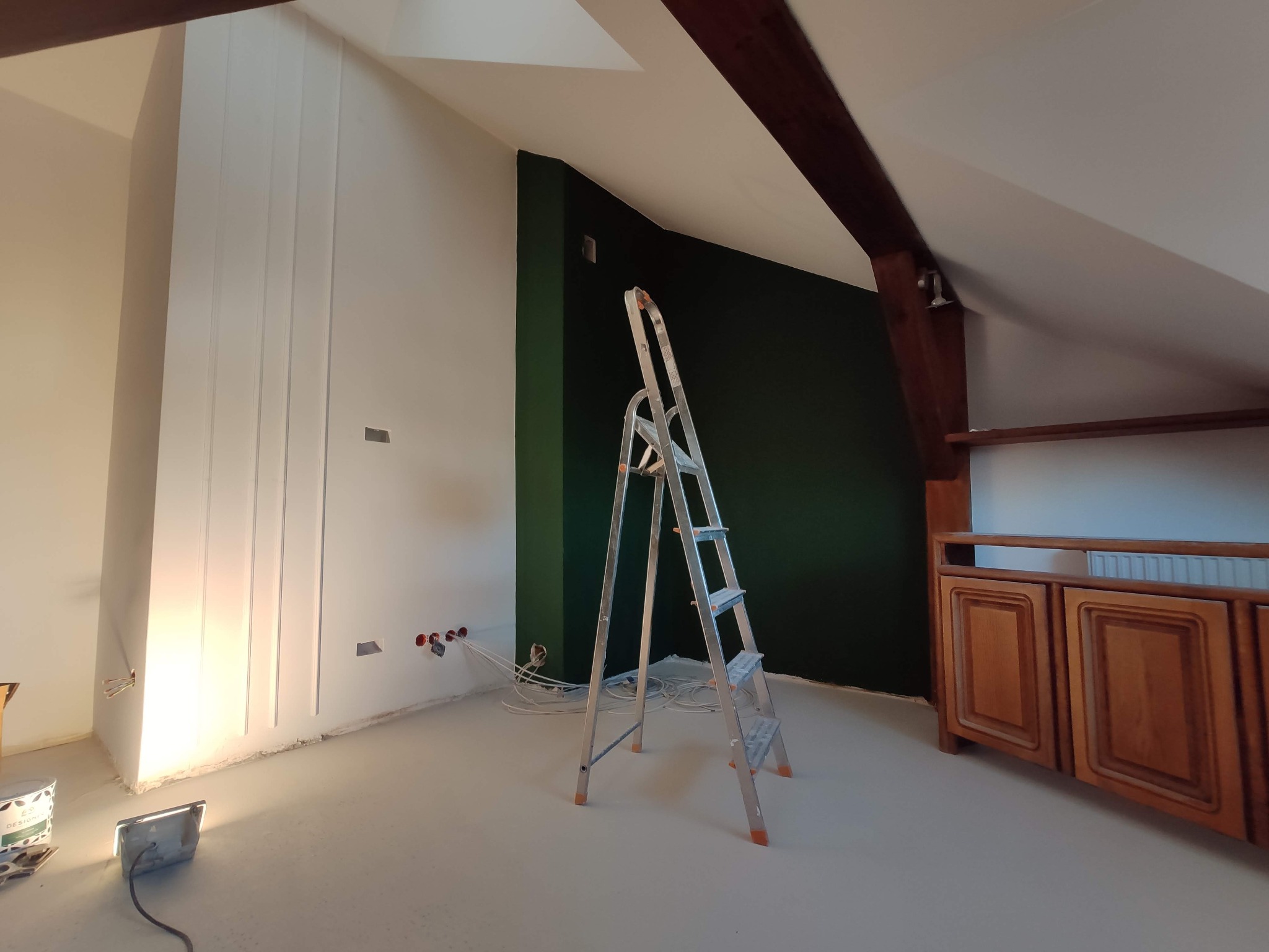 Poddasze w trakcie malowania: ściana pomalowana na ciemnozielono, biała ściana z dekoracyjnymi listwami, aluminiowa drabina, widoczne puszki elektryczne i kable, drewniana belka stropowa