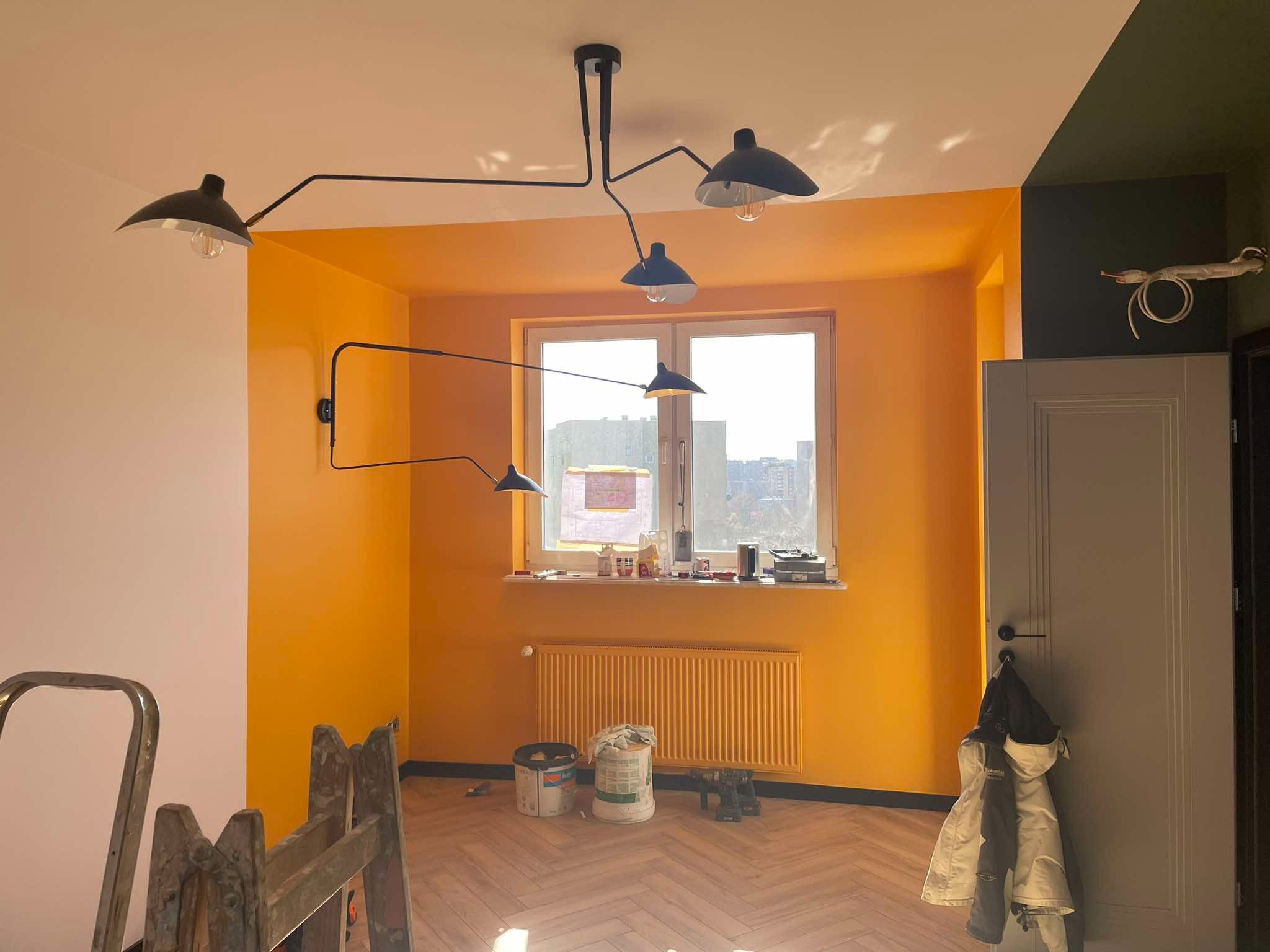 Pokój po malowaniu ścian na żółto-pomarańczowy kolor, widoczne modernistyczne lampy, grzejnik w kolorze ściany, drabina malarska, puszki z farbą, parkiet w jodełkę i szara ściana z drzwiami po...