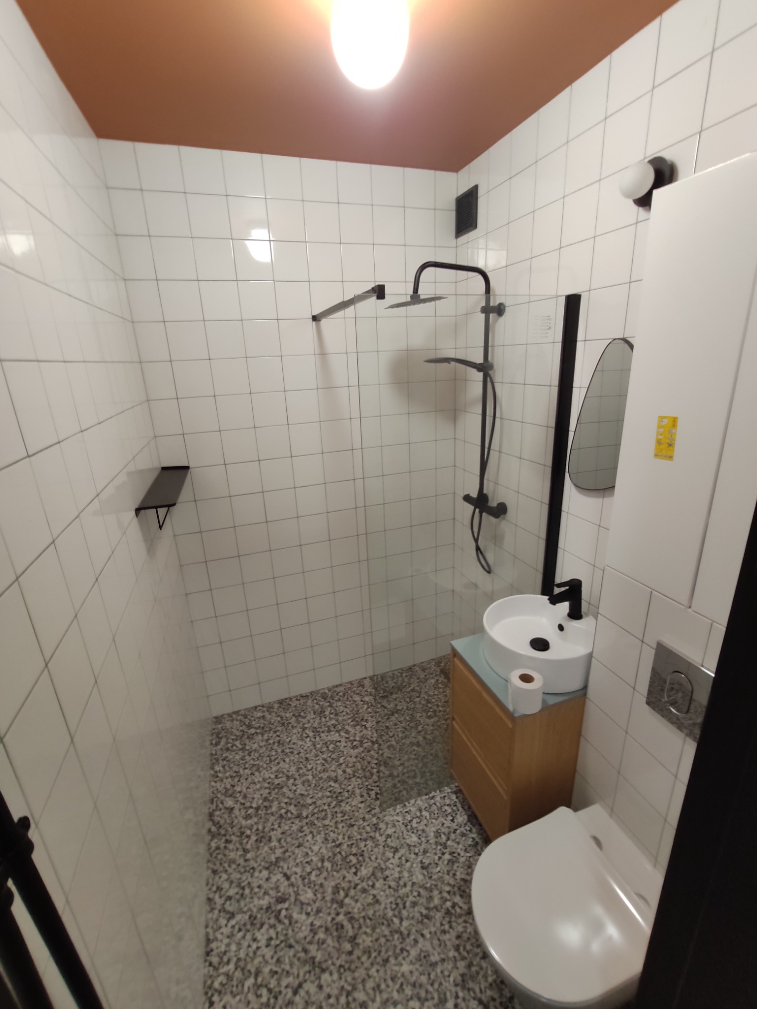 Nowoczesna łazienka z białymi płytkami, deszczownicą w czarnym kolorze, umywalką nablatową na drewnianej szafce i toaletą. Na suficie widoczna pomarańczowa farba.