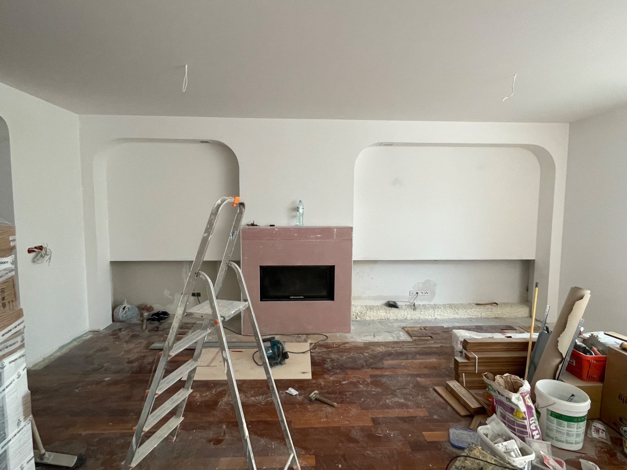 Wnętrze w trakcie remontu w Warszawie: widoczny kominek z obudową w kolorze pudrowego różu, drabina aluminiowa, częściowo położona drewniana podłoga i przygotowane materiały budowlane. Ściany...