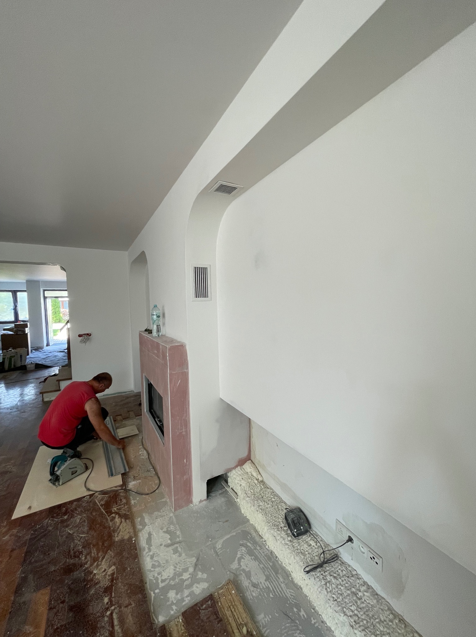 Remont salonu: mężczyzna w czerwonej koszulce przycina płytę pilarką tarczową obok obudowy kominka z różowego gipsu. Widoczne fragmenty starej podłogi, świeżo pomalowane ściany i otwór wentylacyjny.