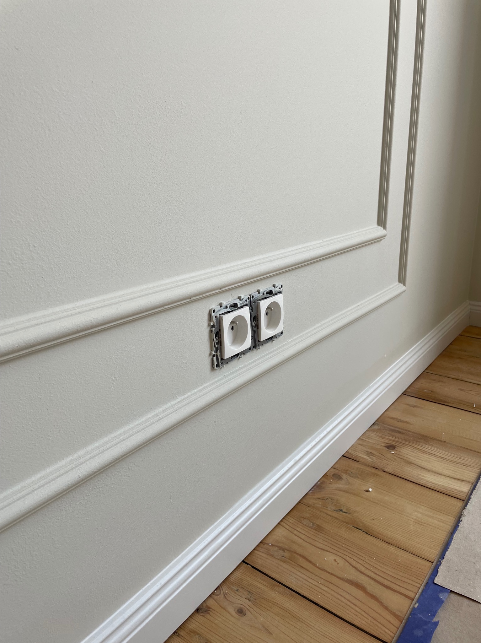 Biała ściana z dekoracyjnymi listwami i zamontowanymi, ale jeszcze niekompletnymi gniazdkami elektrycznymi oraz drewnianą podłogą w trakcie prac wykończeniowych.