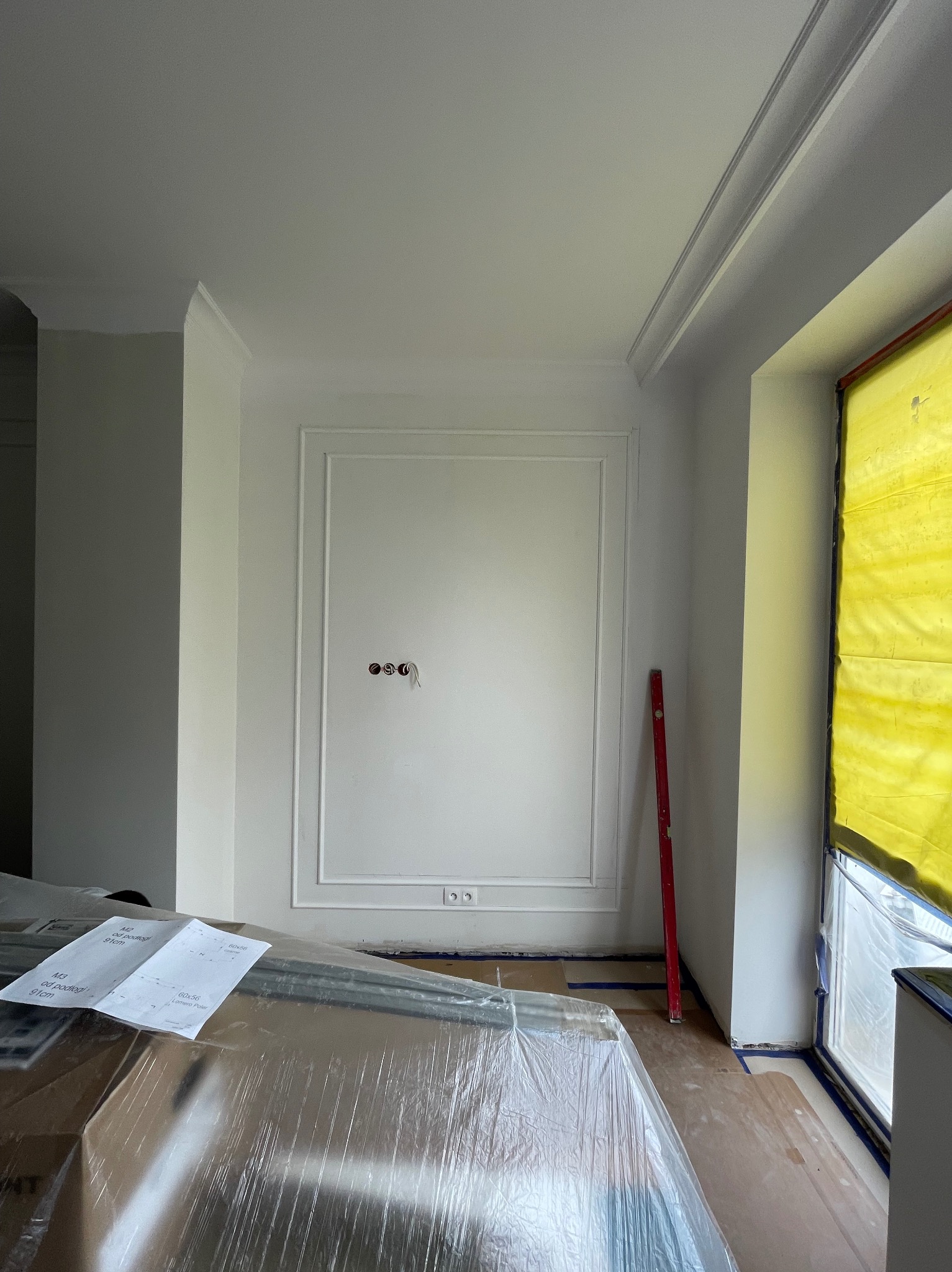 Ściana z dekoracyjną sztukaterią i przygotowanymi punktami elektrycznymi w trakcie prac wykończeniowych, podłoga częściowo zabezpieczona folią, widoczna poziomica. Okno zabezpieczone folią malarską.