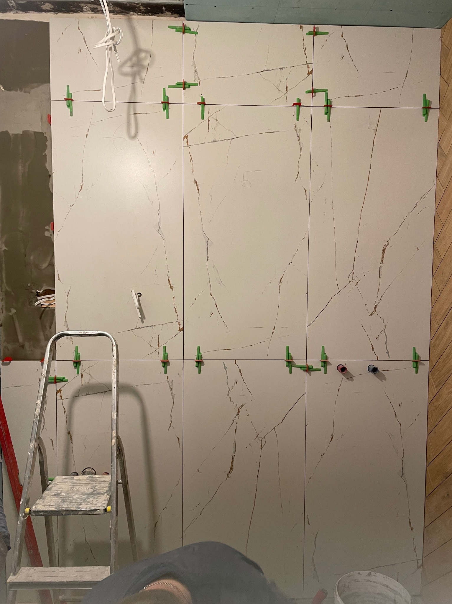 Montaż wielkoformatowych płytek imitujących marmur na ścianie, z użyciem poziomowania krzyżykami, widoczna drabina i fragment instalacji wodnej.