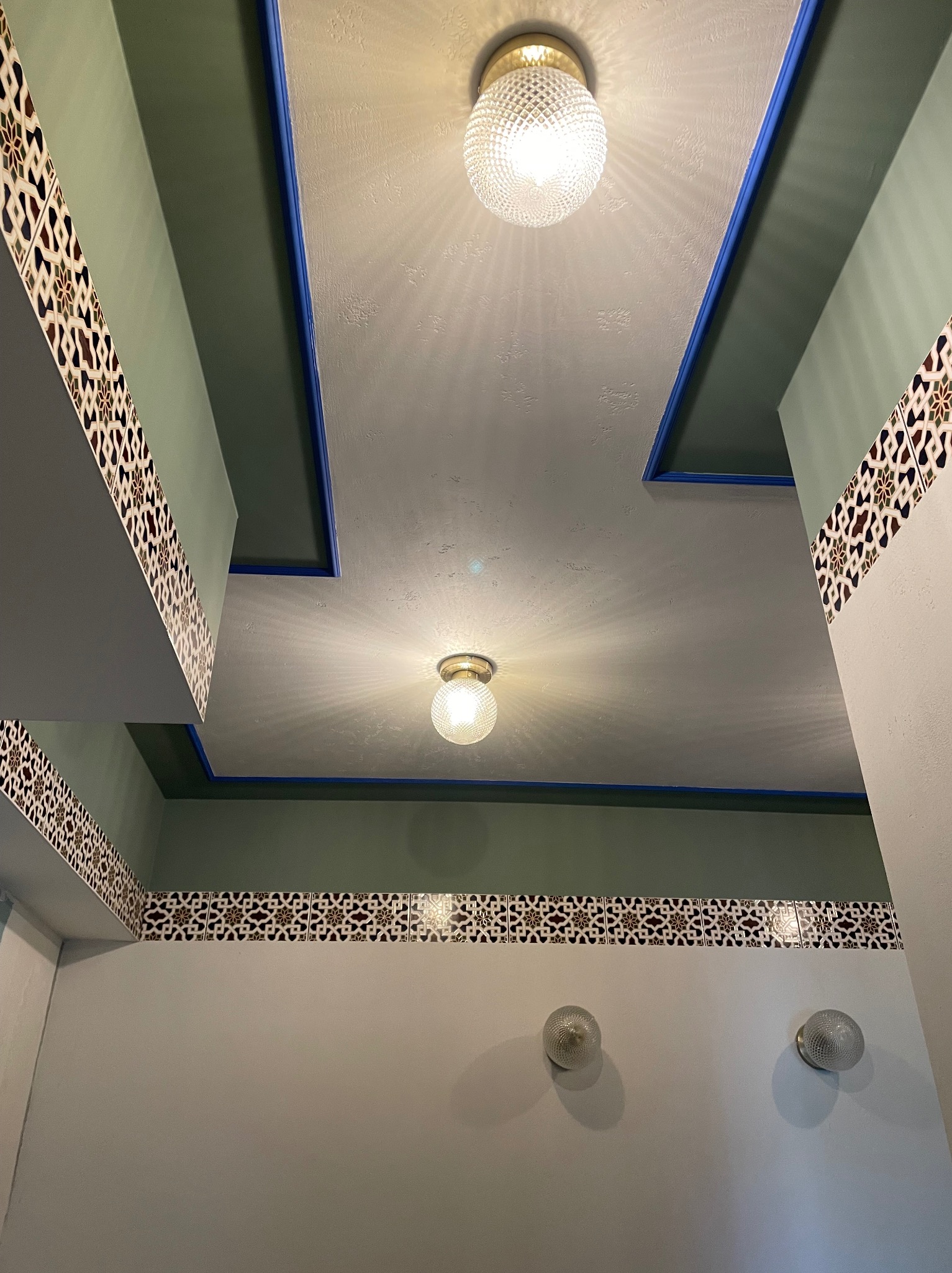 Wnętrze łazienki w Warszawie z geometrycznym wzorem płytek na ścianie i suficie, oświetlone dwoma okrągłymi lampami ściennymi i dwoma sufitowymi z kloszem o fakturze diamentu.