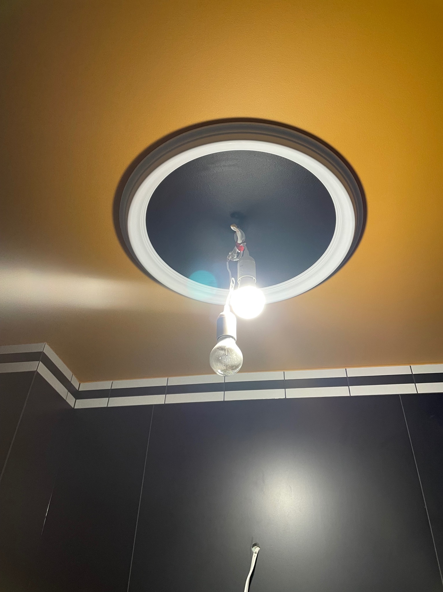 Żółty sufit z czarnym, okrągłym elementem dekoracyjnym i prowizorycznym oświetleniem w postaci dwóch żarówek na kablu, widoczne fragmenty czarnych płytek z białym paskiem