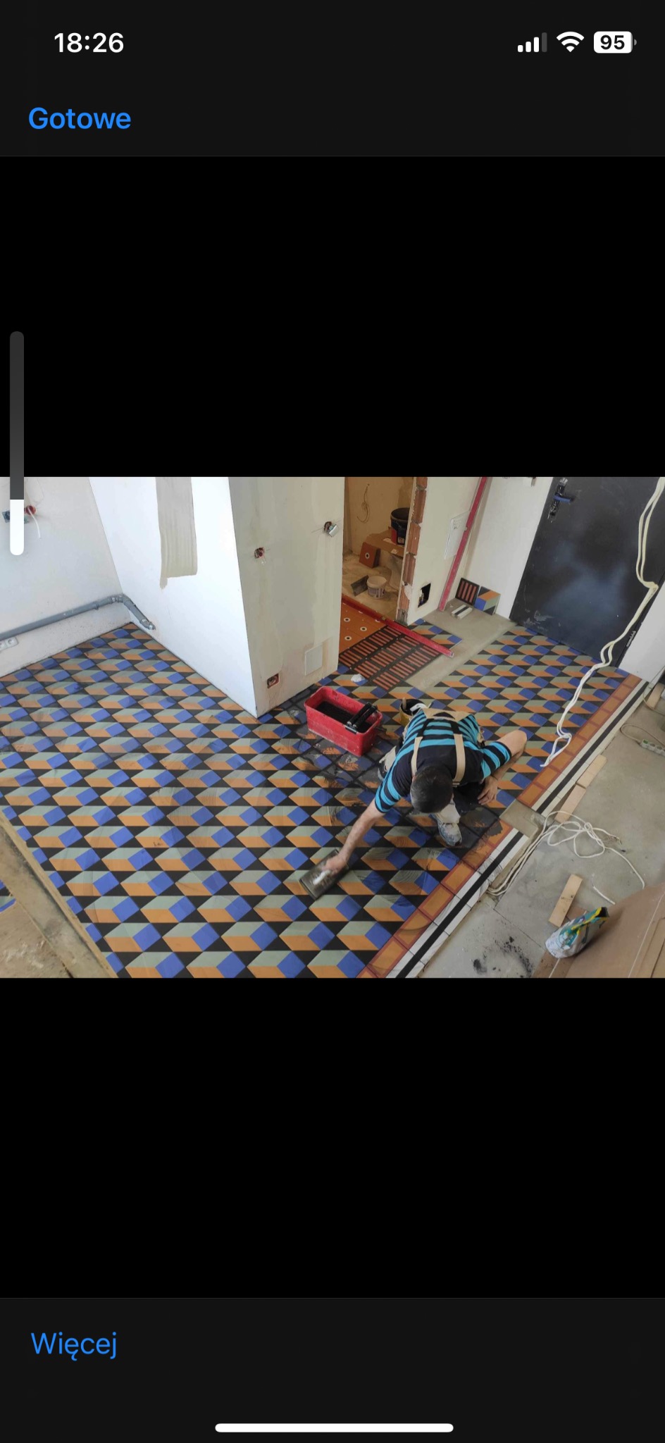 Układanie wzorzystej podłogi w pomieszczeniu, widoczny pracownik rozprowadzający fugę na nowo położonych płytkach z geometrycznym wzorem 3D, czerwone wiadro z narzędziami obok.