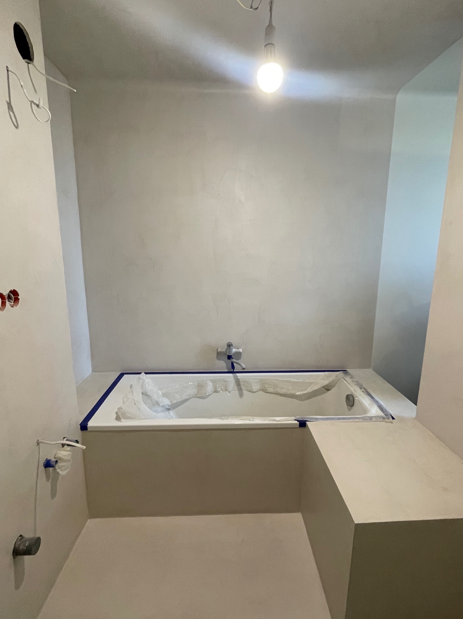 Łazienka w trakcie remontu: wanna zabezpieczona folią i taśmą malarską, surowe ściany przygotowane do malowania, widoczne instalacje elektryczne i hydrauliczne.
