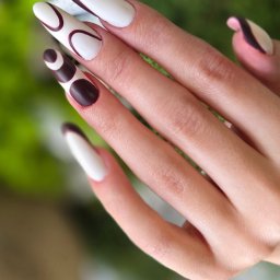 JOWIN JOANNA WINKIEL - Dłoń z długimi, białymi paznokciami ozdobionymi brązowymi, geometrycznymi wzorami na tle zieleni. Nietypowy, nowoczesny design manicure.