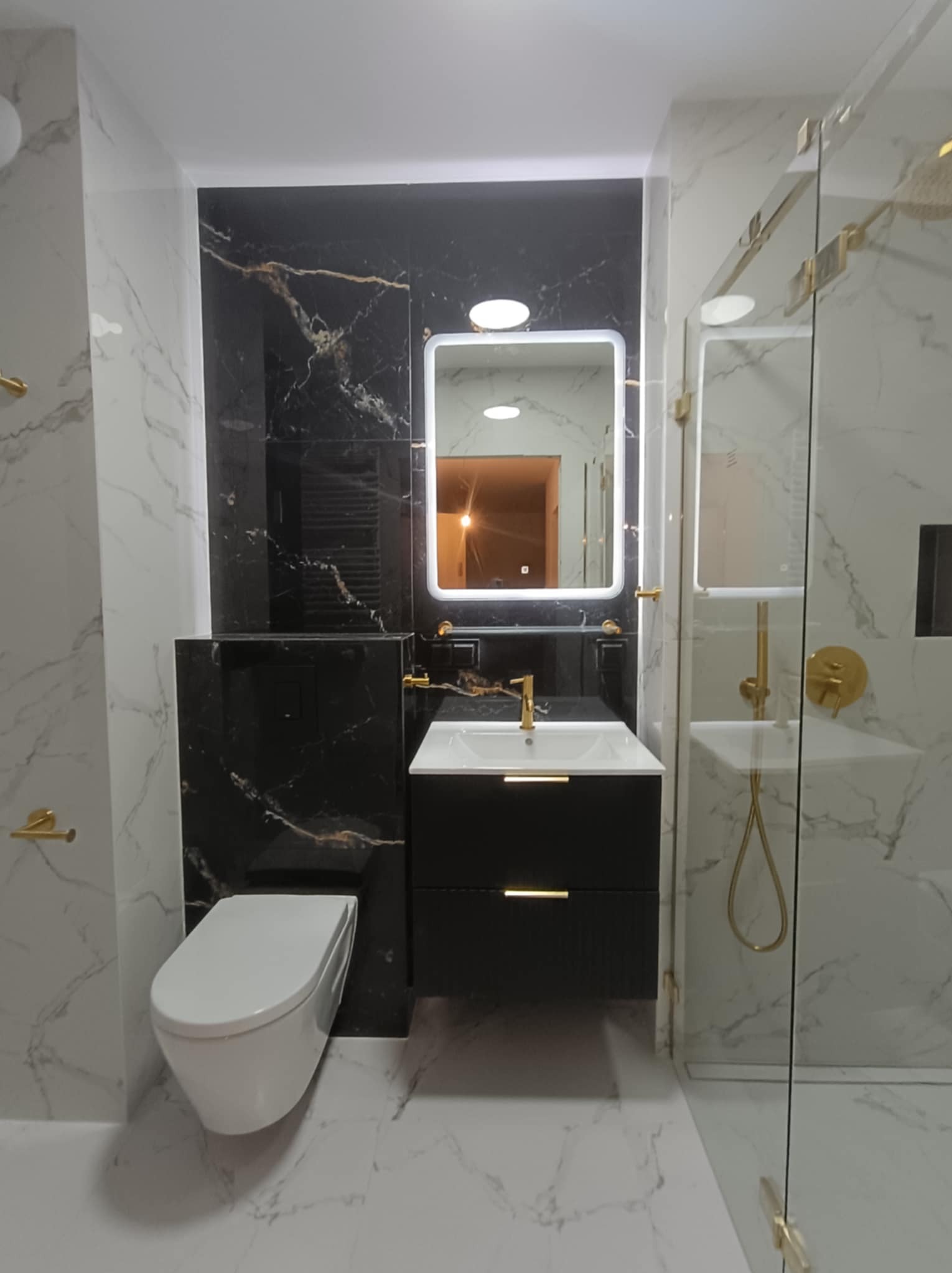 Elegancka łazienka po remoncie: biało-czarne marmurowe płytki, złote akcenty armatury, podświetlane lustro i kabina prysznicowa ze złotymi okuciami.