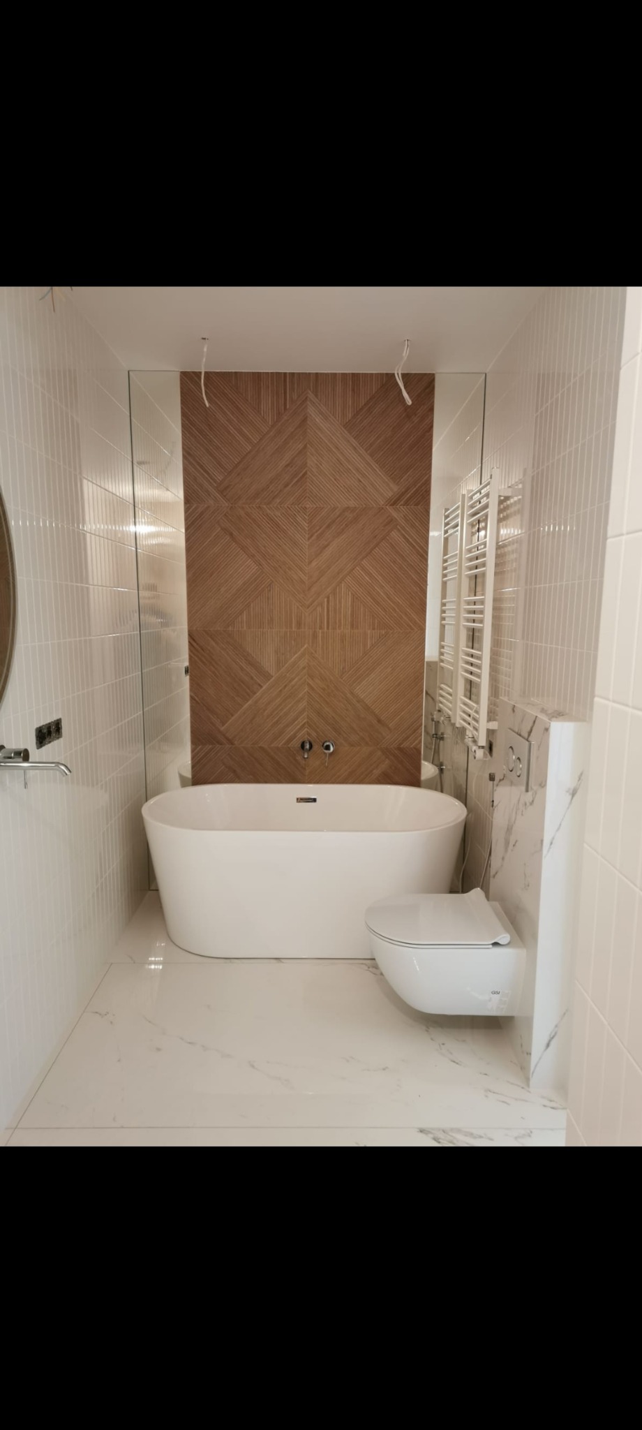 Nowoczesna łazienka z wolnostojącą wanną, toaletą podwieszaną i dekoracyjną ścianą z drewnianych paneli ułożonych w geometryczny wzór. Białe płytki na ścianach i podłodze.