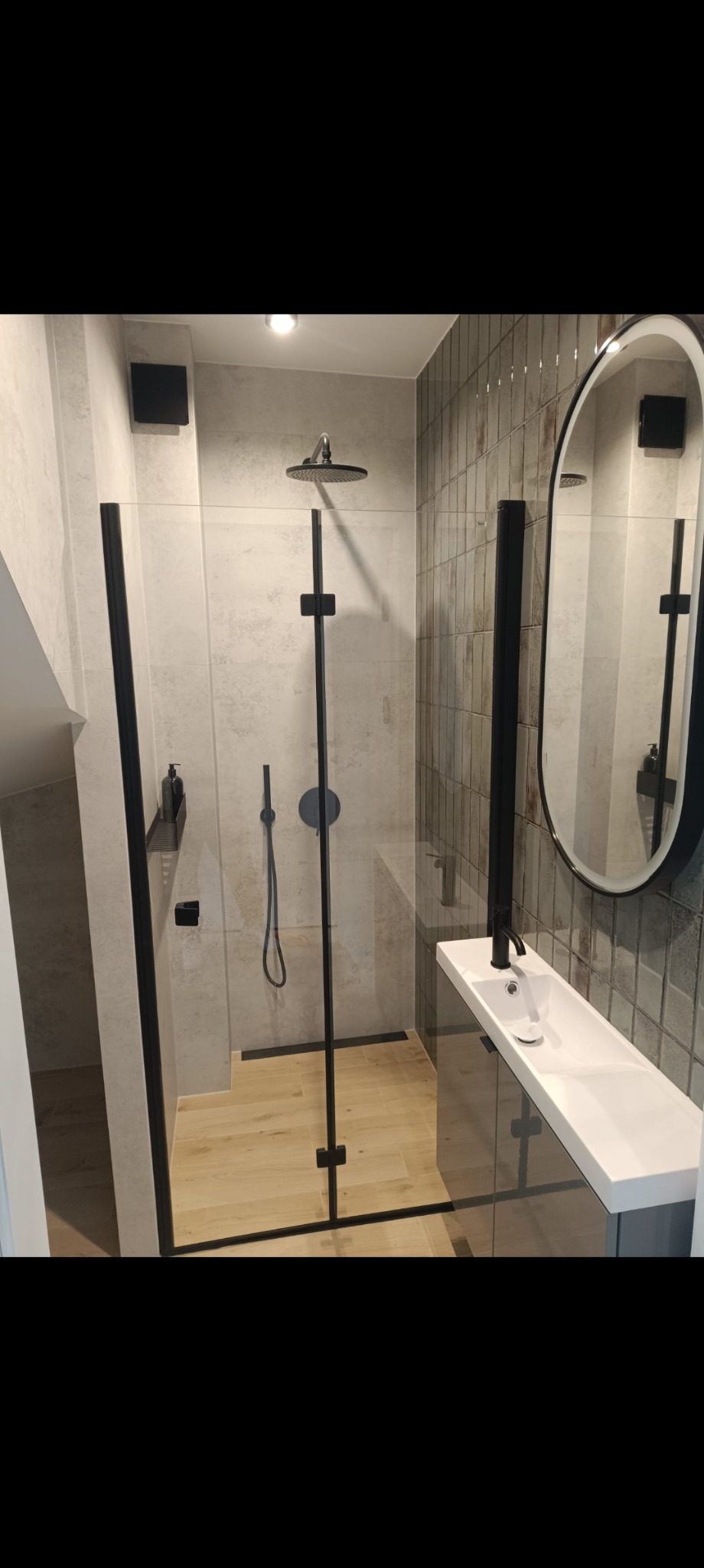 Nowoczesna łazienka z kabiną typu walk-in z czarnymi profilami, drewnopodobną podłogą i podłużną umywalką z lustrem w czarnej ramie.