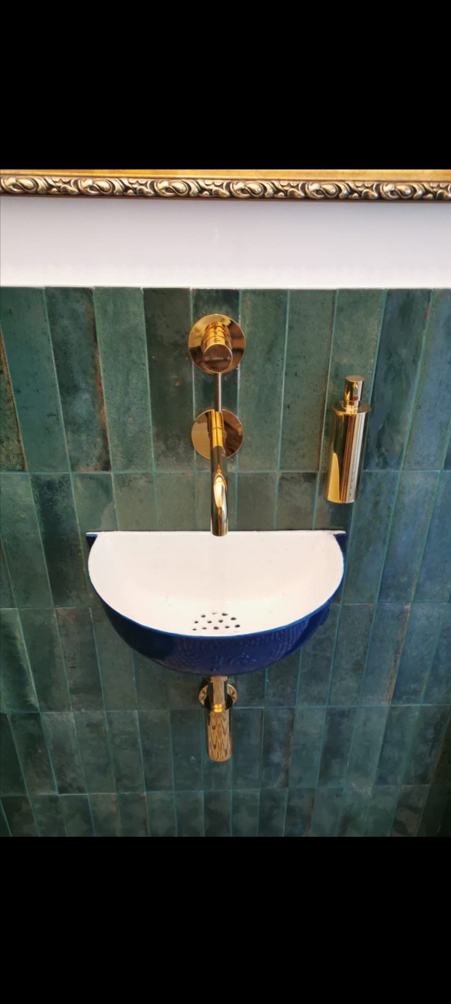 Nowoczesna umywalka w kolorze białym i niebieskim z złotą armaturą na tle ściany wyłożonej zielonymi, podłużnymi płytkami w łazience.