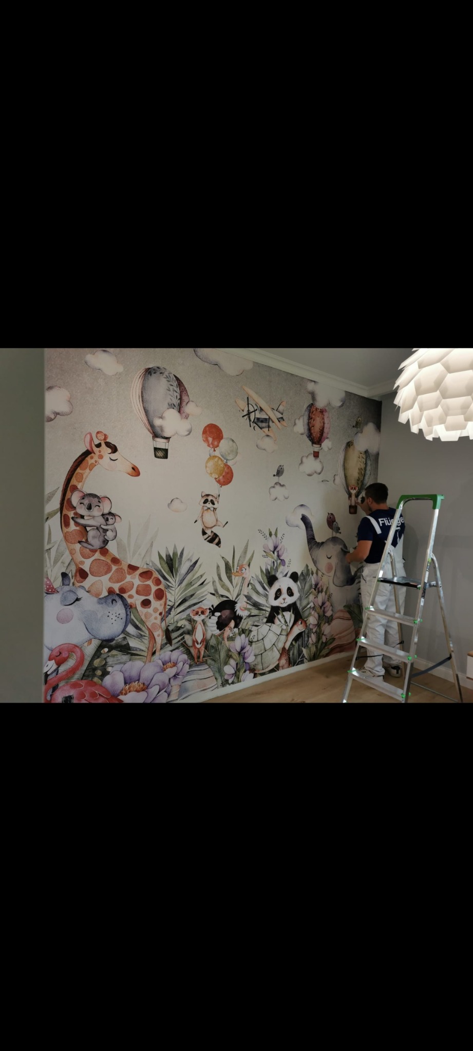 Pracownik na drabinie montuje tapetę z motywem zwierząt i balonów w pokoju dziecięcym. Widoczne detale tapety i narzędzia pracy.