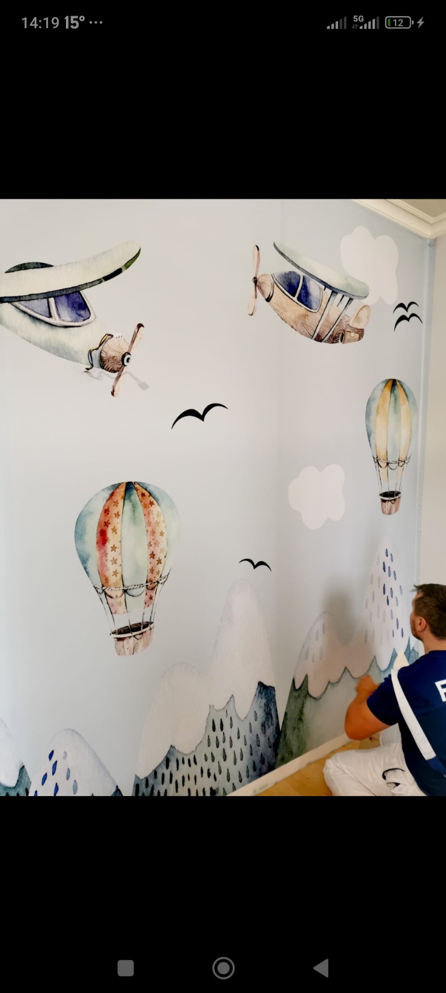 Człowiek nakleja na ścianę w pokoju dziecięcym tapetę z motywem gór, samolotów i balonów na tle jasnego, pomalowanego na niebiesko pokoju.