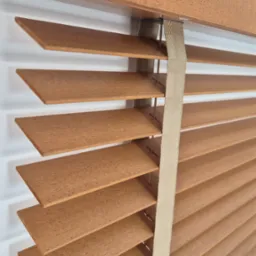 Zamknięte drewniane żaluzje w ciepłym odcieniu brązu, z widoczną strukturą drewna i beżową taśmą podtrzymującą, zamontowane na białej ścianie.