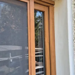 Brązowa moskitiera przesuwna na oknie, widok z zewnątrz, siatka zabezpieczająca przed owadami, z bliska, widoczne odbicie otoczenia w szybie.