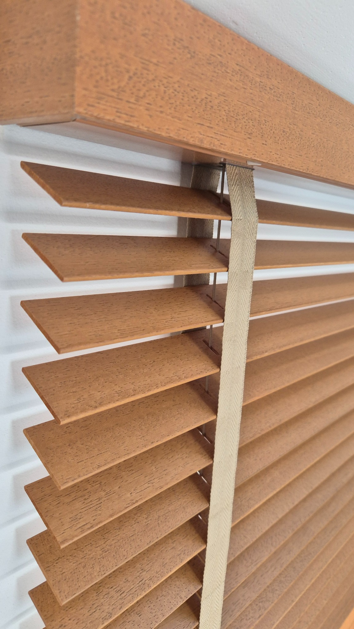 Zamknięte drewniane żaluzje w ciepłym odcieniu brązu, z widoczną strukturą drewna i beżową taśmą podtrzymującą, zamontowane na białej ścianie.