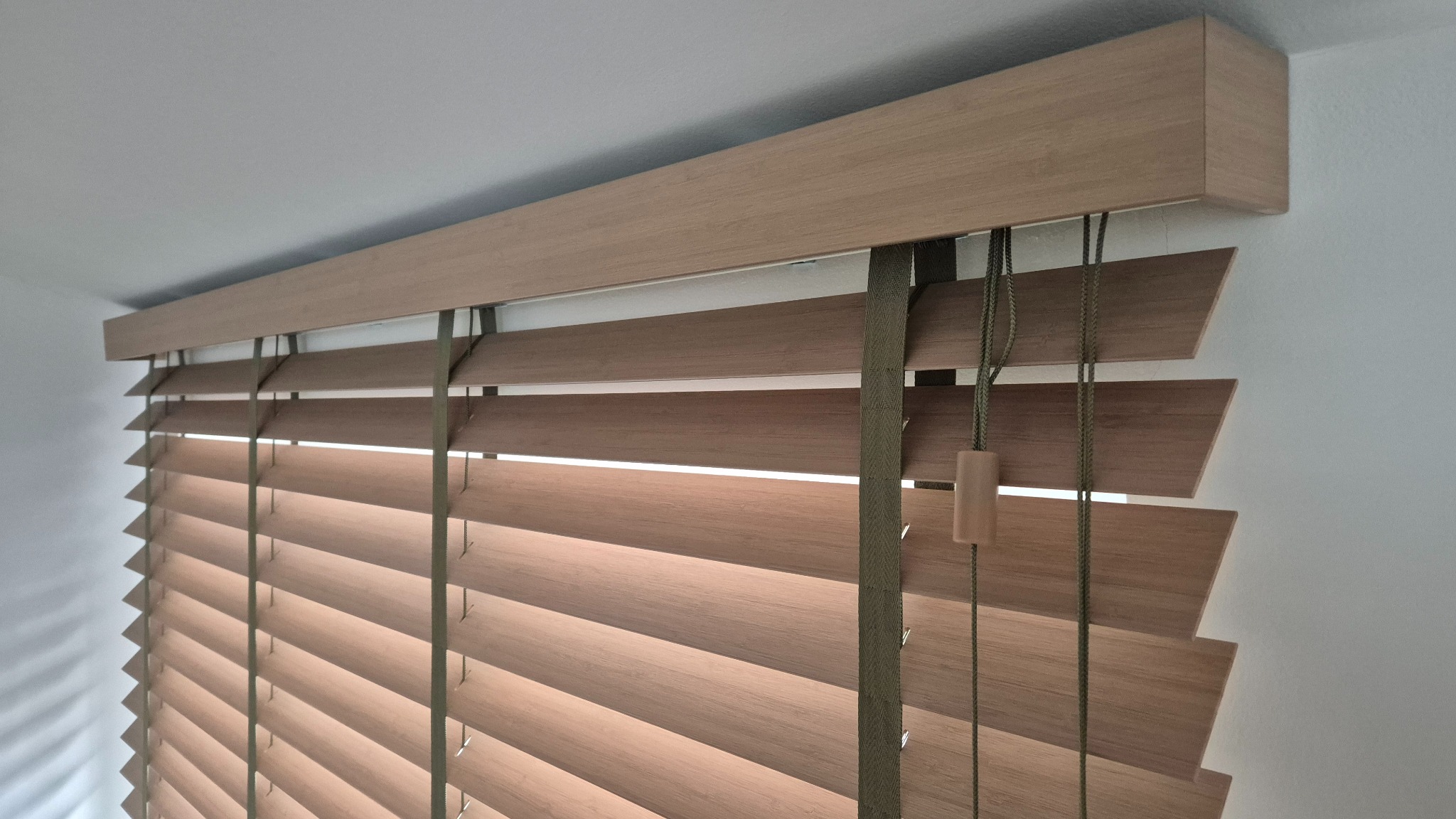 Zamknięte drewniane żaluzje w odcieniach brązu, z widocznym mechanizmem sznurkowym i taśmami, zamontowane przy białej ścianie. Delikatne światło prześwituje między lamelami.