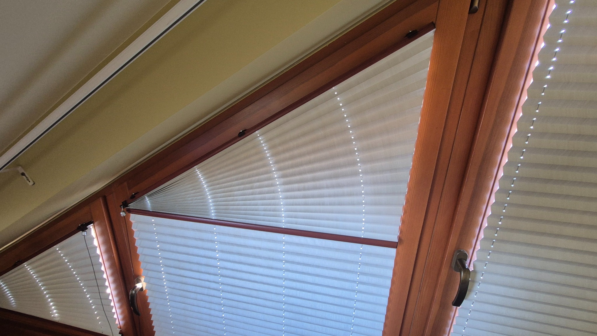 Jasne, plisowane osłony okienne w drewnianej ramie okna na poddaszu, z widocznym uchwytem okiennym i fragmentem skośnego sufitu.