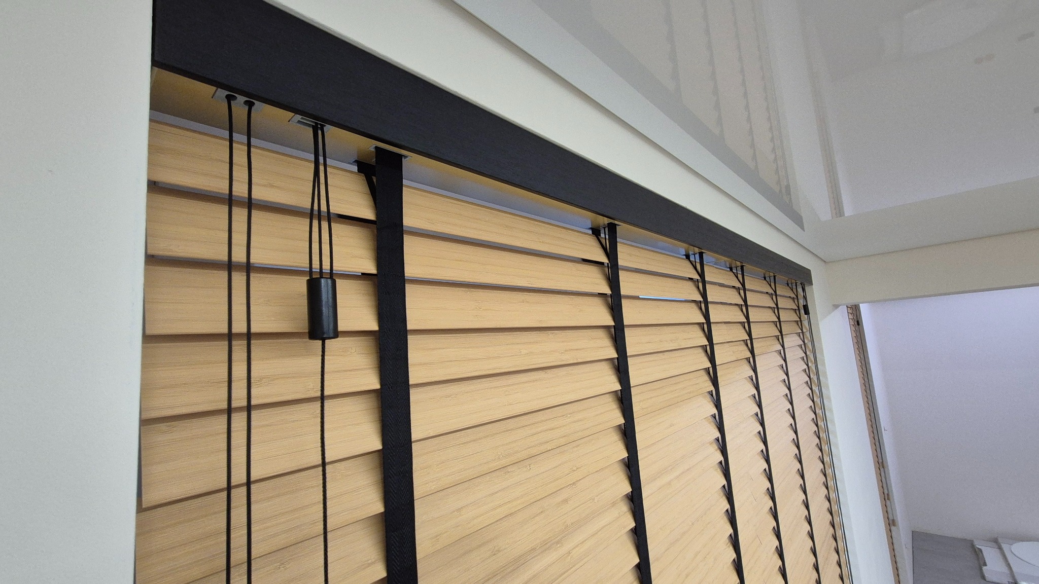 Jasne, drewniane lamele żaluzji z czarnymi elementami montażowymi i sznurkami, zamontowane w oknie z białymi ścianami. Ujęcie z bliska, widoczne detale.