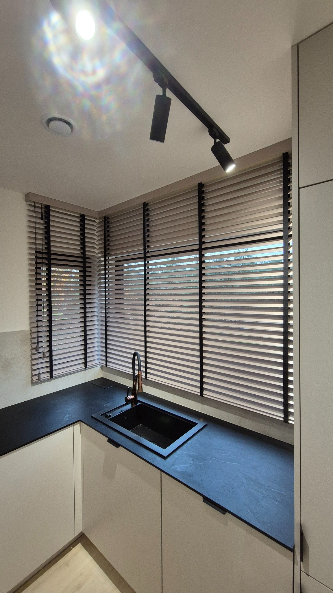 Jasne żaluzje z czarnymi taśmami w nowoczesnej kuchni. Widoczne okno, czarny zlew i bateria. Oświetlenie sufitowe punktowe.