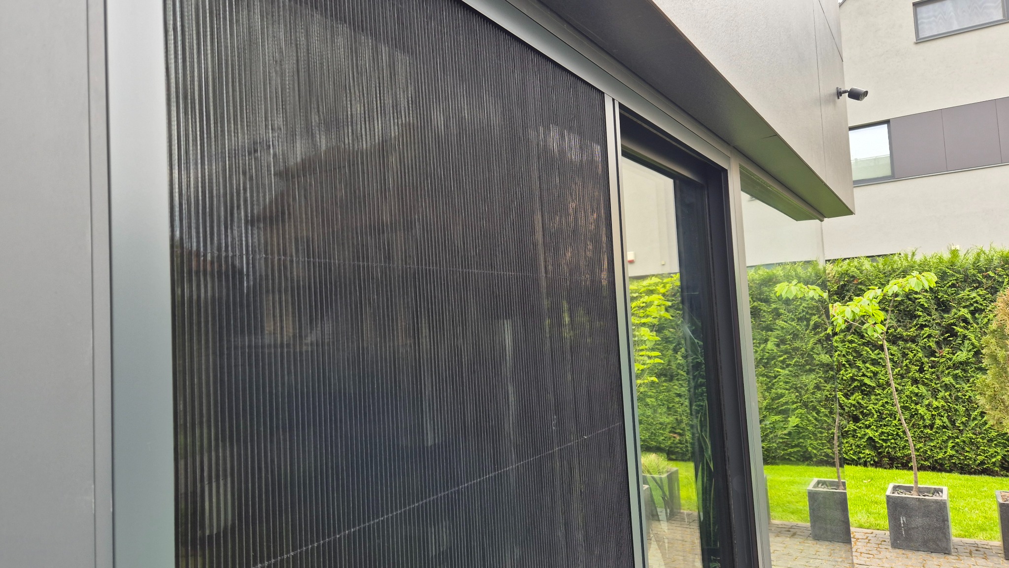 Zamknięta, czarna, plisowana moskitiera okienna w nowoczesnym budynku. Widok z zewnątrz, zieleń w tle odbija się w szybie.