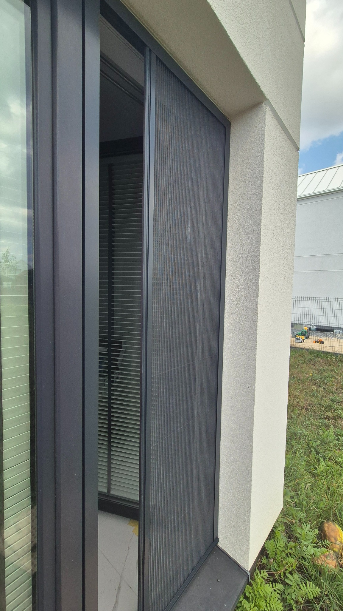 Zewnętrzna, grafitowa plisa okienna zamontowana na nowoczesnym oknie balkonowym. Widoczny fragment elewacji budynku i trawnika. Ujęcie z bliska.