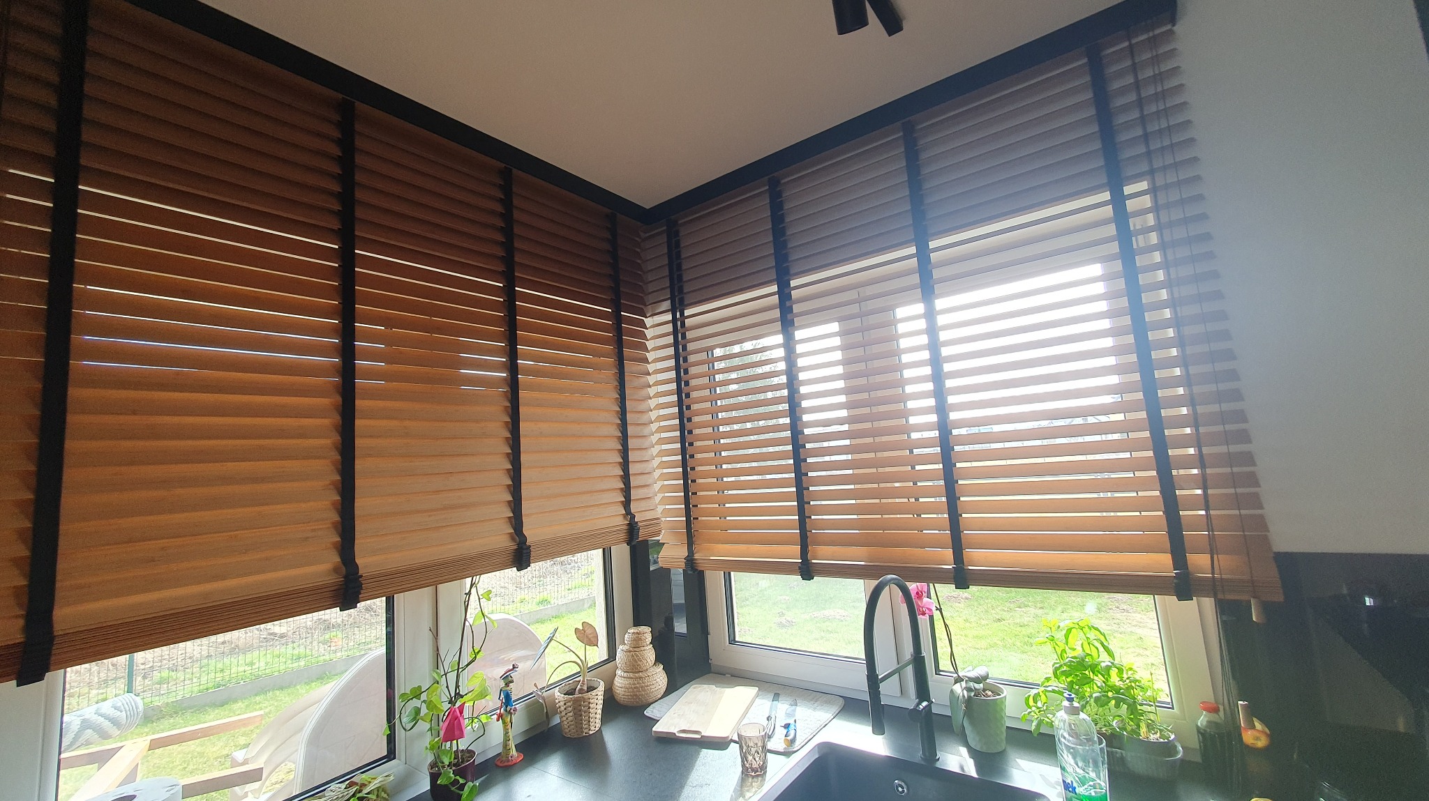 Jasne żaluzje drewniane z czarnymi taśmami w kuchni, częściowo zasłonięte okna z widokiem na zieleń. Blat kuchenny z dekoracjami i roślinami.