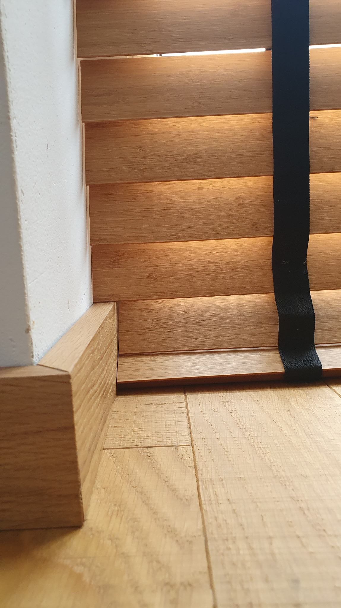 Fragment drewnianych żaluzji z czarnym paskiem, widoczne przy ścianie i podłodze z paneli. Ujęcie z dołu, ukazujące detale wykończenia.