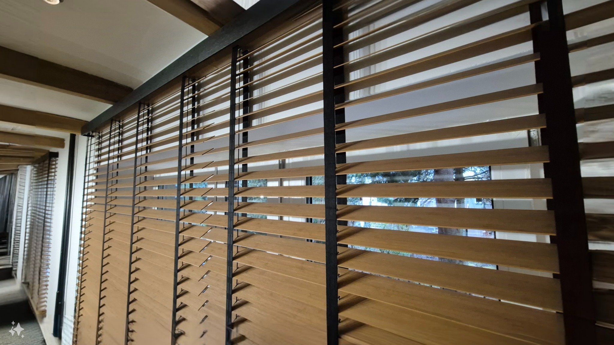 Zamknięte drewniane żaluzje z czarnymi akcentami, widoczne przez szpary drzewa na zewnątrz. Ustawione równolegle, tworzą wzór linii w ciepłym oświetleniu wnętrza.