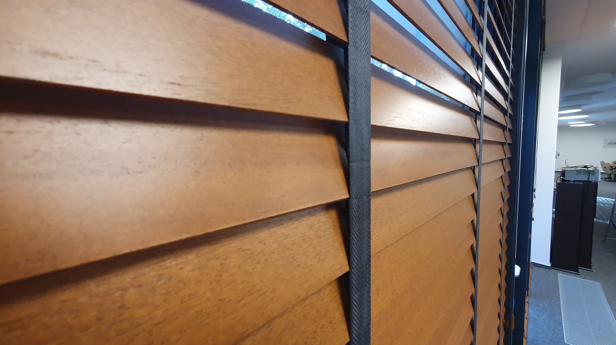 Brązowe żaluzje drewniane z ciemnymi taśmami na tle jasnego pomieszczenia biurowego, widok z boku, podkreślona faktura drewna i detale wykończenia.