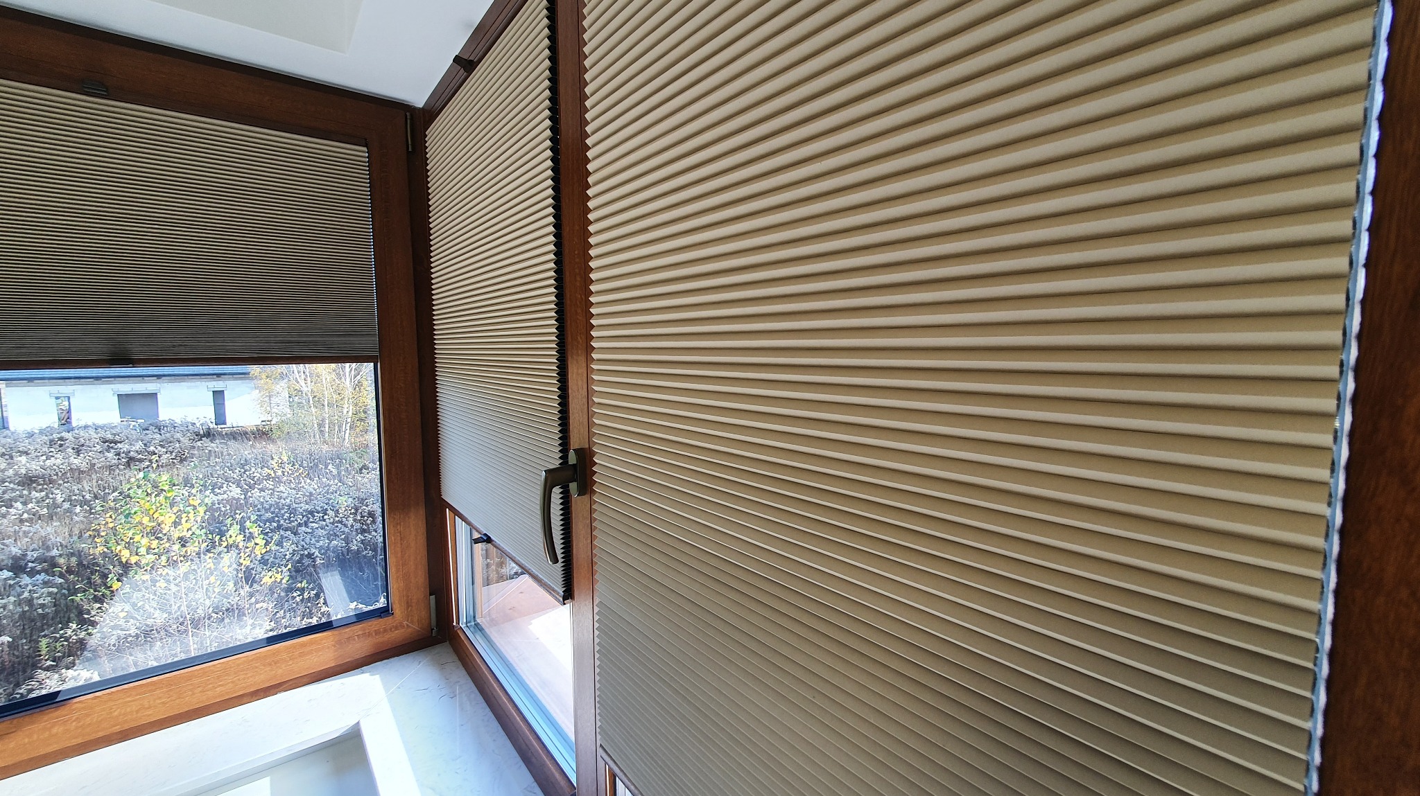 Beżowe plisy okienne w drewnianej ramie, widok z bliska. Jedna plisa częściowo zasłonięta, druga odsłonięta, ukazująca widok na zewnątrz.