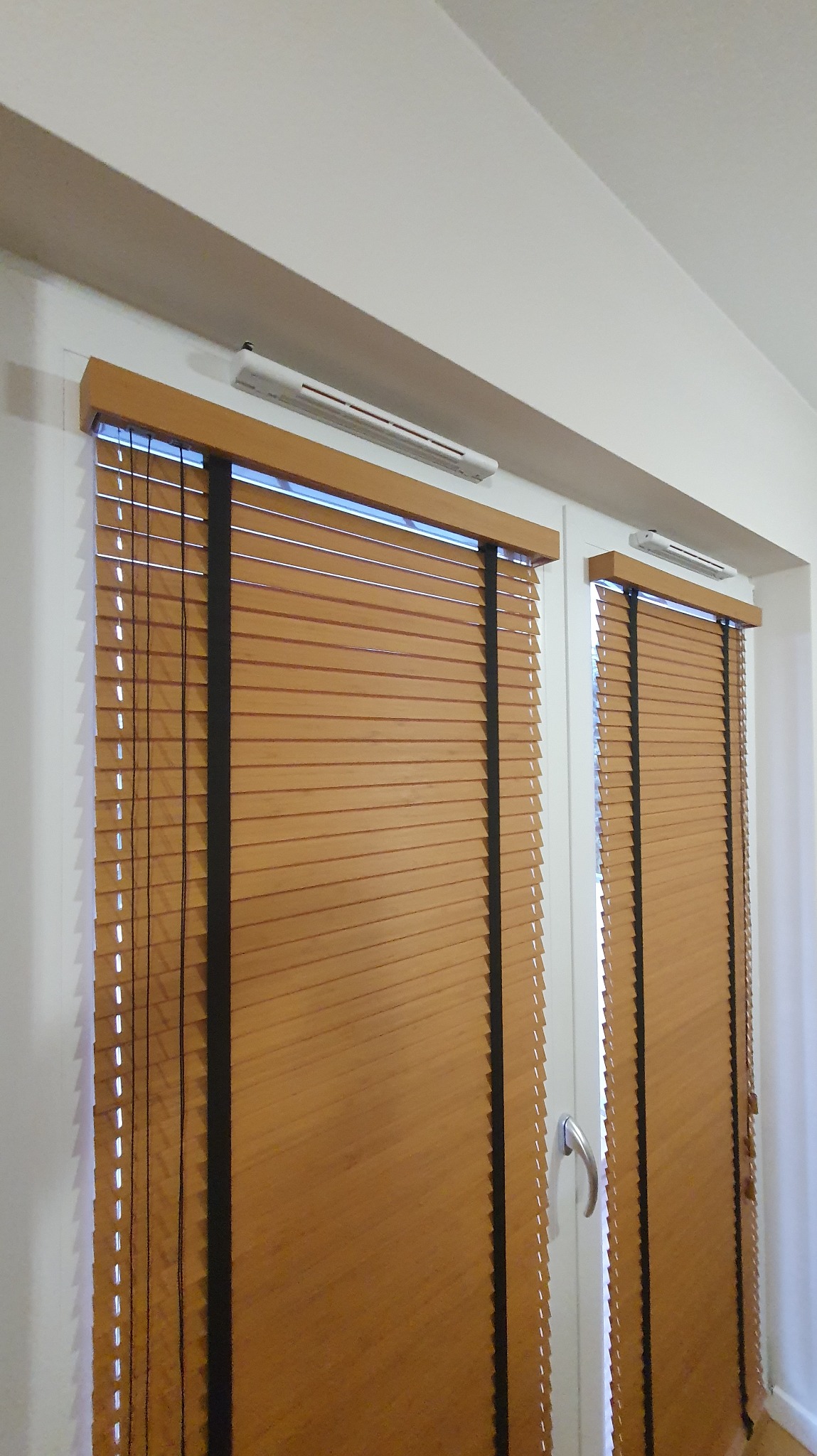 Zamknięte, poziome żaluzje drewniane w kolorze miodowego brązu, z czarnymi taśmami, zamontowane na białych oknach. Widoczne nawiewniki okienne.